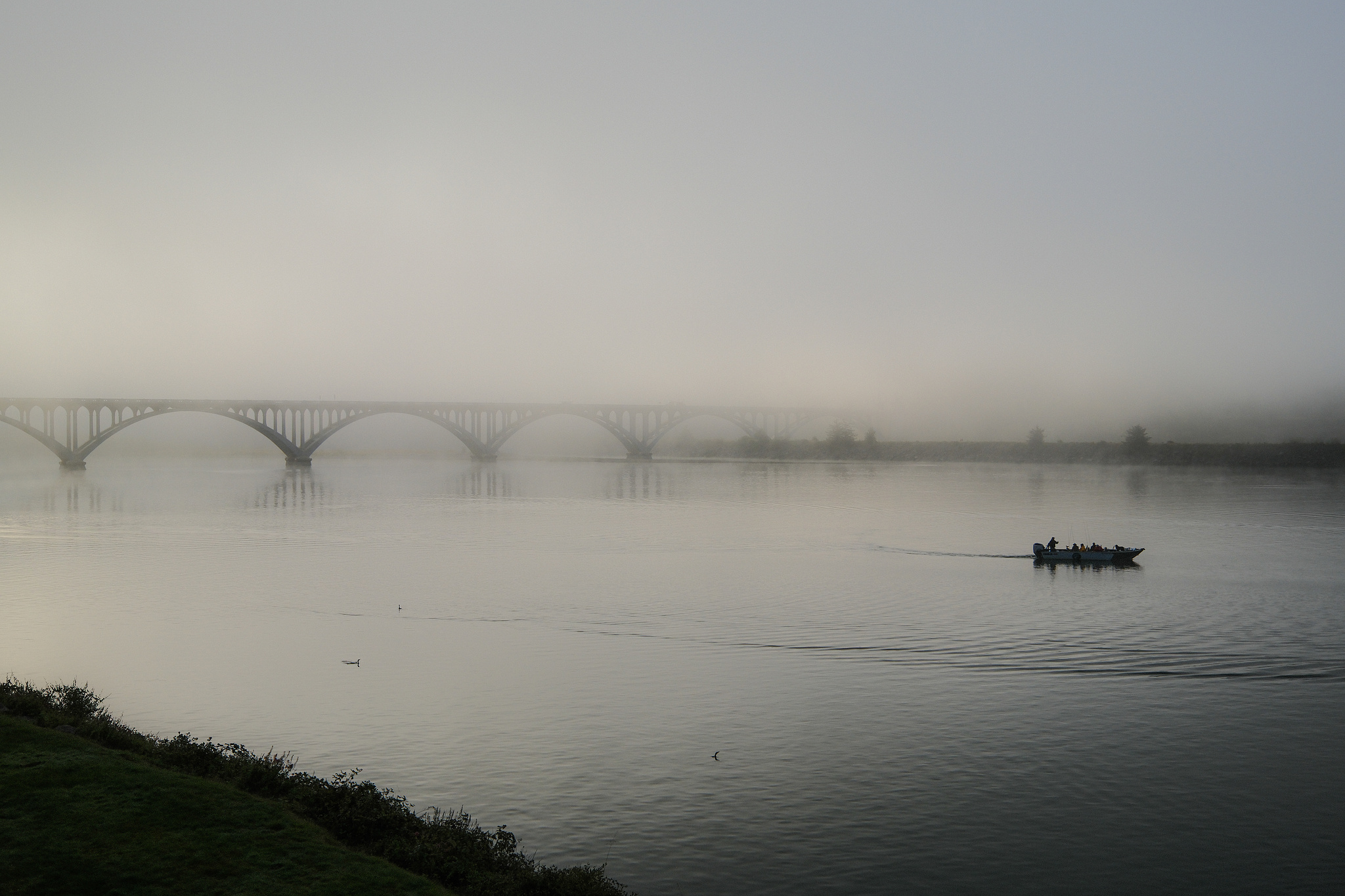 A fishing boat heads toward sea, a fog-covered bridge in the background