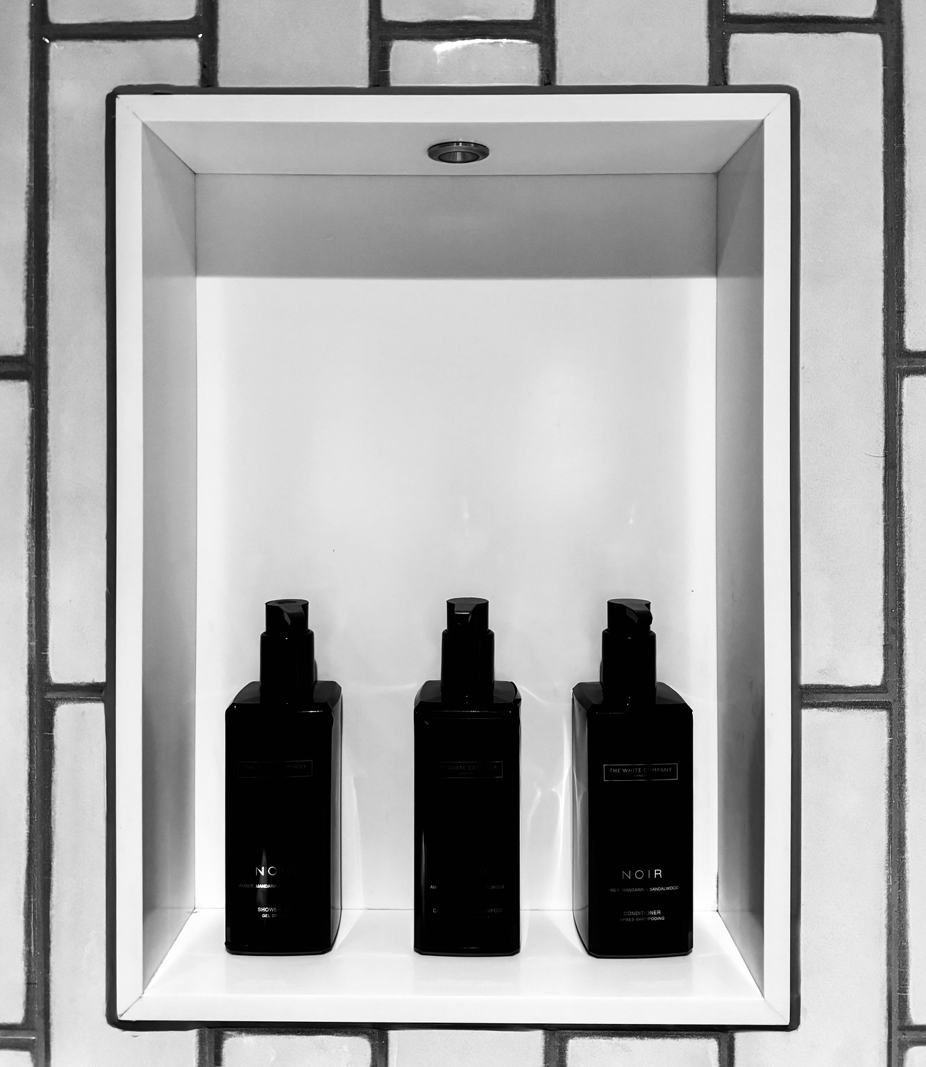 Three black bottles on a white shelf against a white tiled wall.