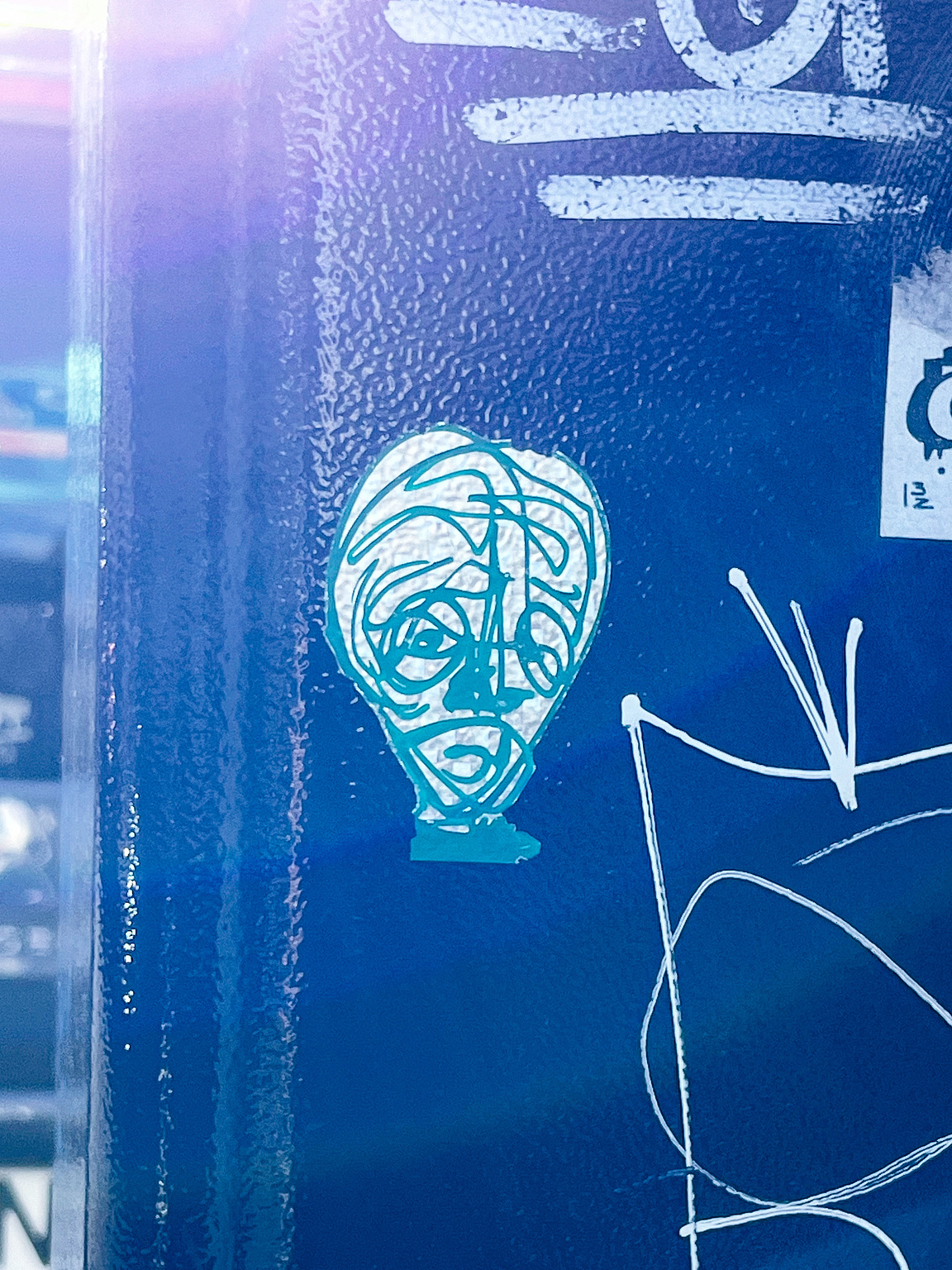 A blue-toned sticker depicts a stylized, abstract human head with tangled lines, affixed to a textured surface with other graffiti.