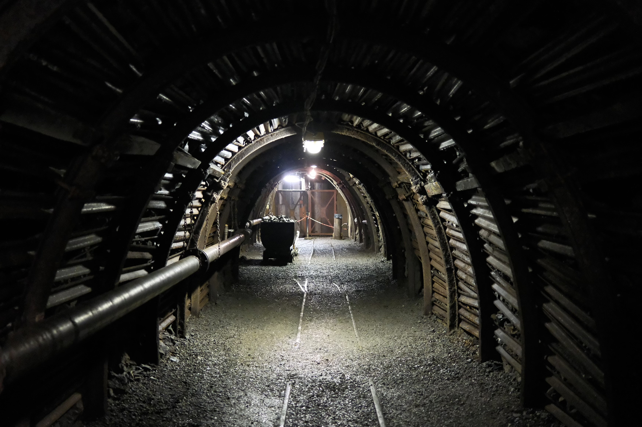 Donkere mijnschacht met boogvormige stalen ondersteuning, spoorlijnen op de grond en een transportkar in de verte, spaarzaam verlicht door mijnlampen.