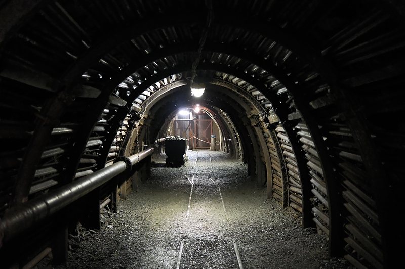 Donkere mijnschacht met boogvormige stalen ondersteuning, spoorlijnen op de grond en een transportkar in de verte, spaarzaam verlicht door mijnlampen.