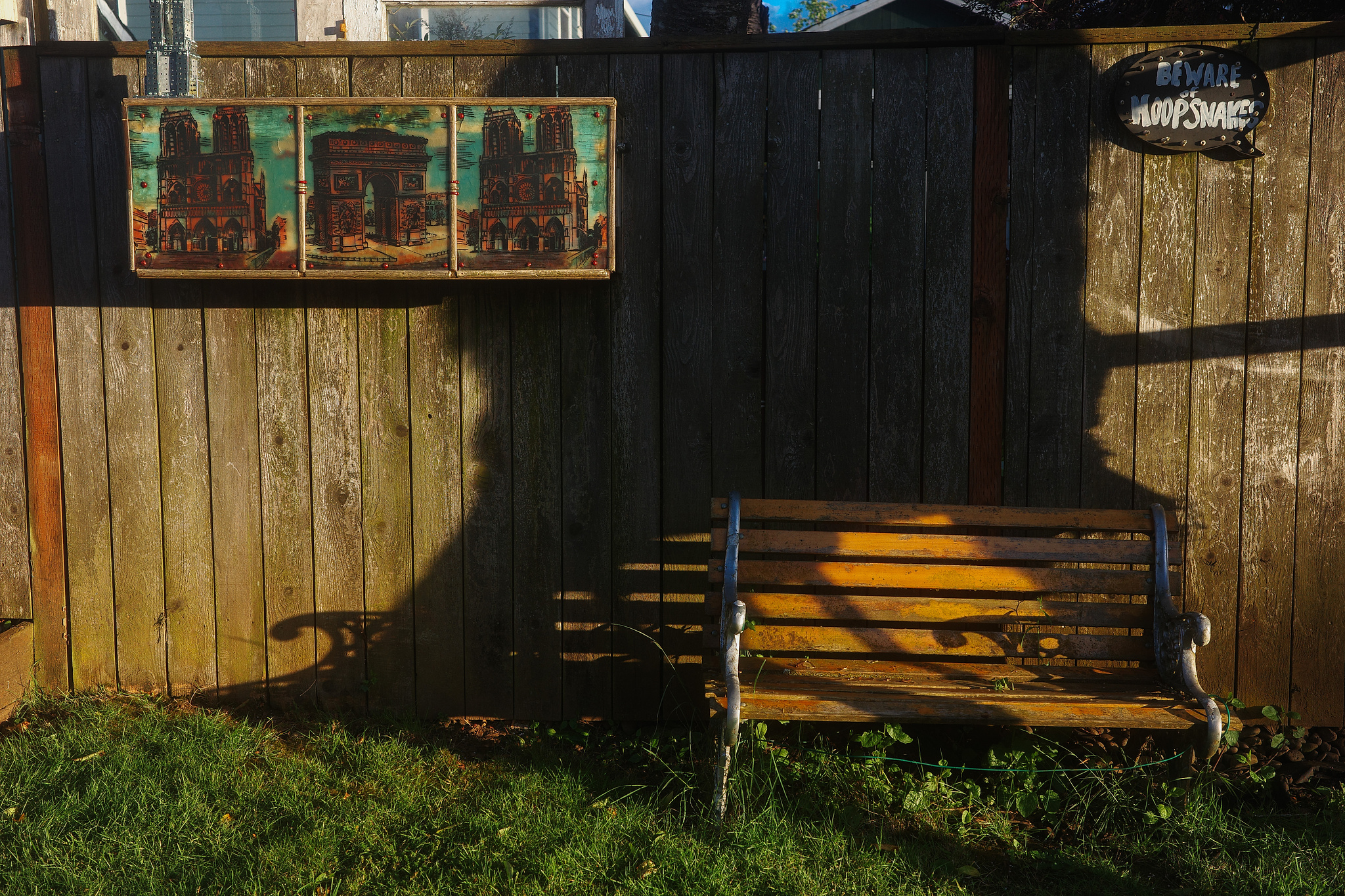 A brown park bench against a fence with a triptych of painted images of Paris in a frame. 