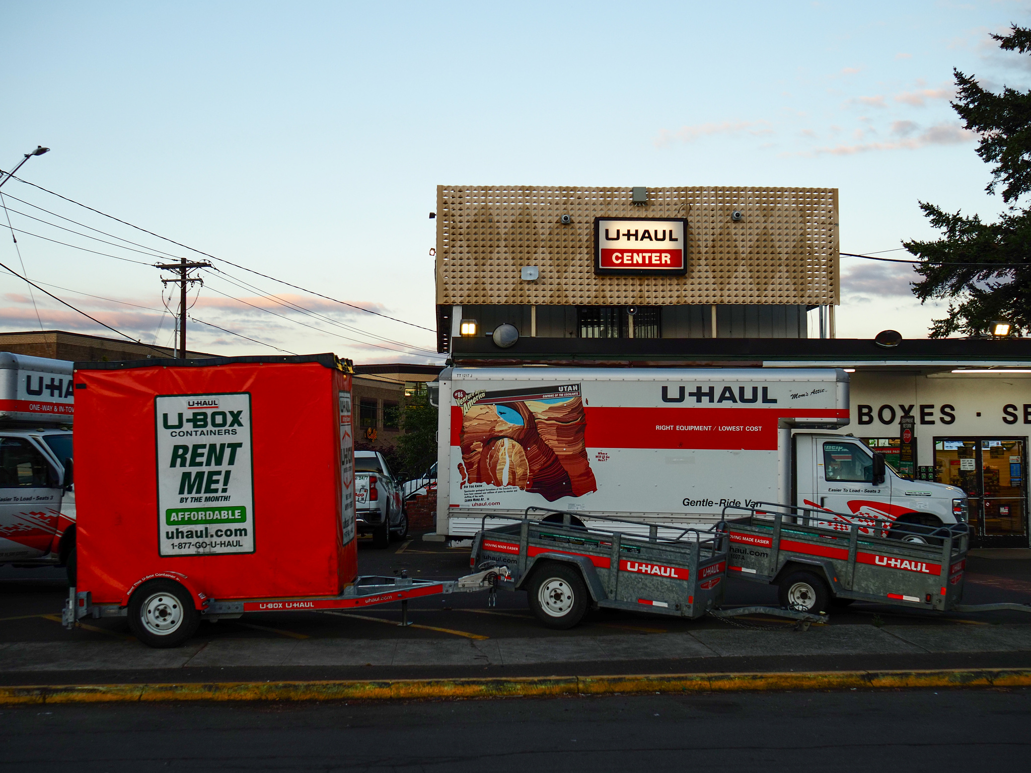 A bright orange U-Haul trailer and U-Haul van in front of a building with a glowing "U-Haul Center" sign at sunset. 