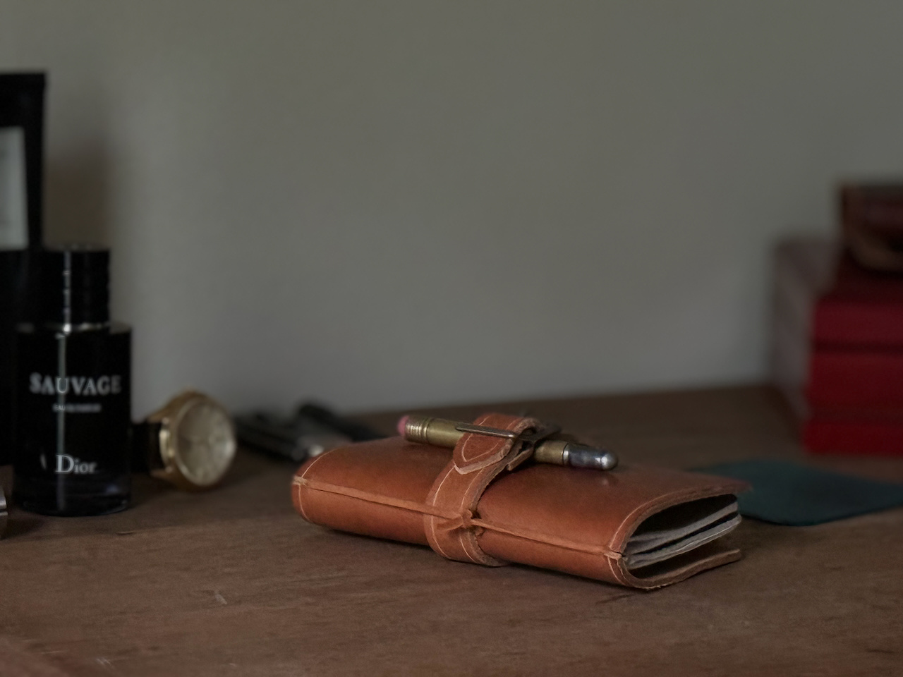 A small leather notebook or wallet resting on a wooden surface, with a pen clipped onto it. The background includes a black bottle of Sauvage cologne, a watch, and some other indistinct objects, creating a sophisticated and elegant atmosphere.