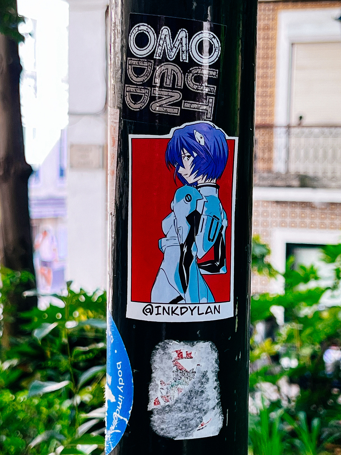 Stickers on a pole, featuring a blue-haired animated character, layered among others; urban background. Text includes "@INKYDYLAN."