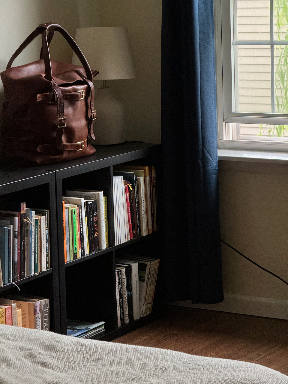 A leather bag is placed on a black bookshelf beside a window, partially covered by a curtain. The bookshelf is filled with books, and a table lamp is seen on top. The scene suggests a cozy, well-organized room with natural light streaming in through the window.