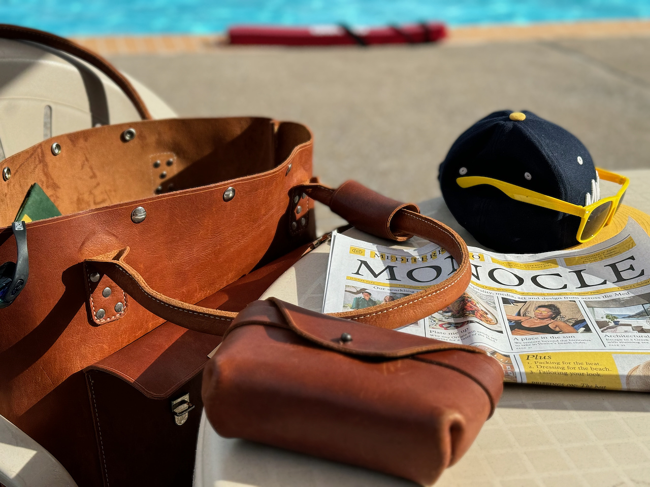 A scene by the poolside with a leather bag opened on a chair, alongside a newspaper, sunglasses, and a baseball cap. The pool water is visible in the background, creating a relaxed, vacation-like atmosphere.