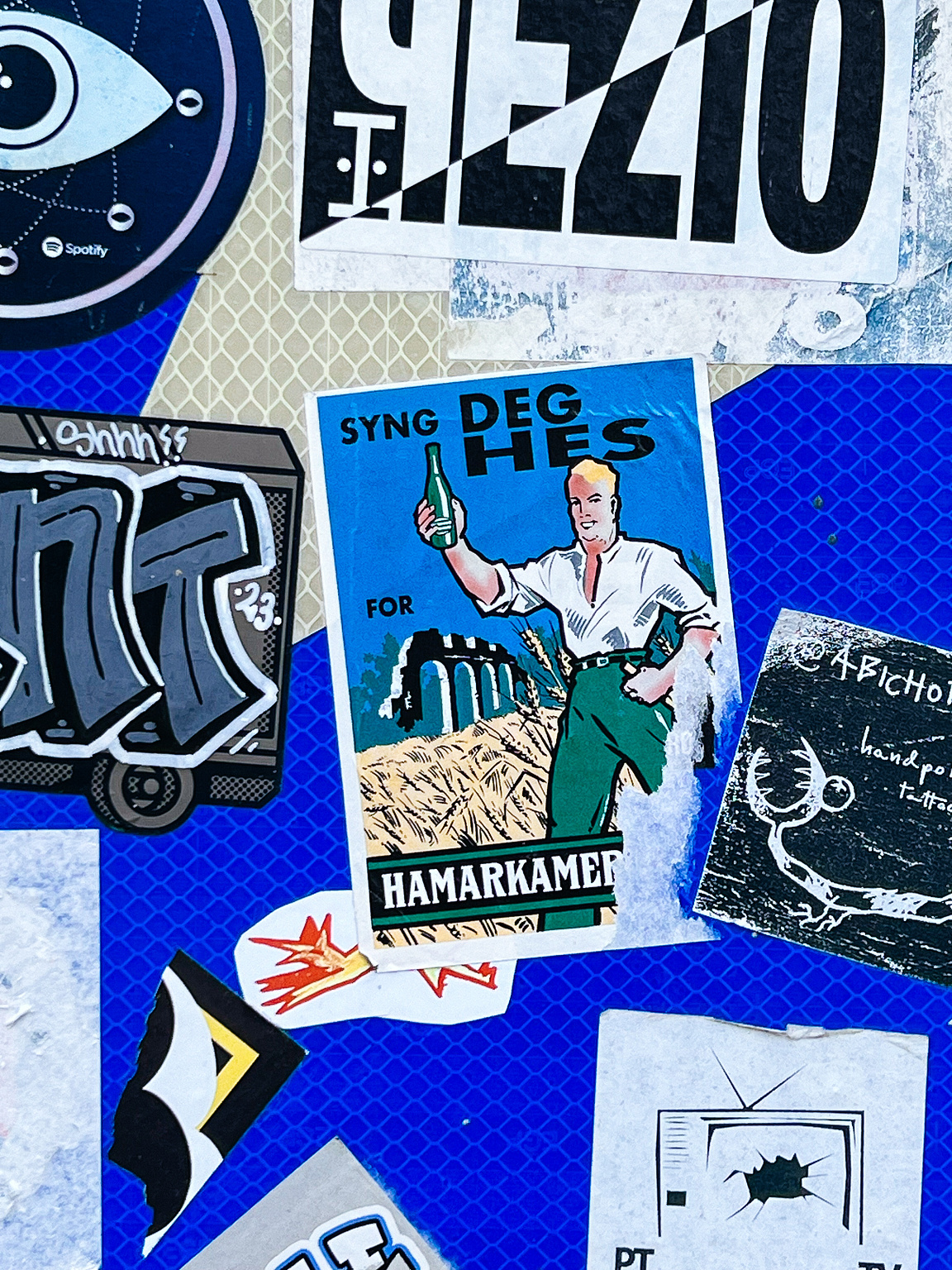 A collage of assorted stickers on a surface; one depicts a man holding a beverage with text "SYNG DEG HES FOR HAMARKAMER".