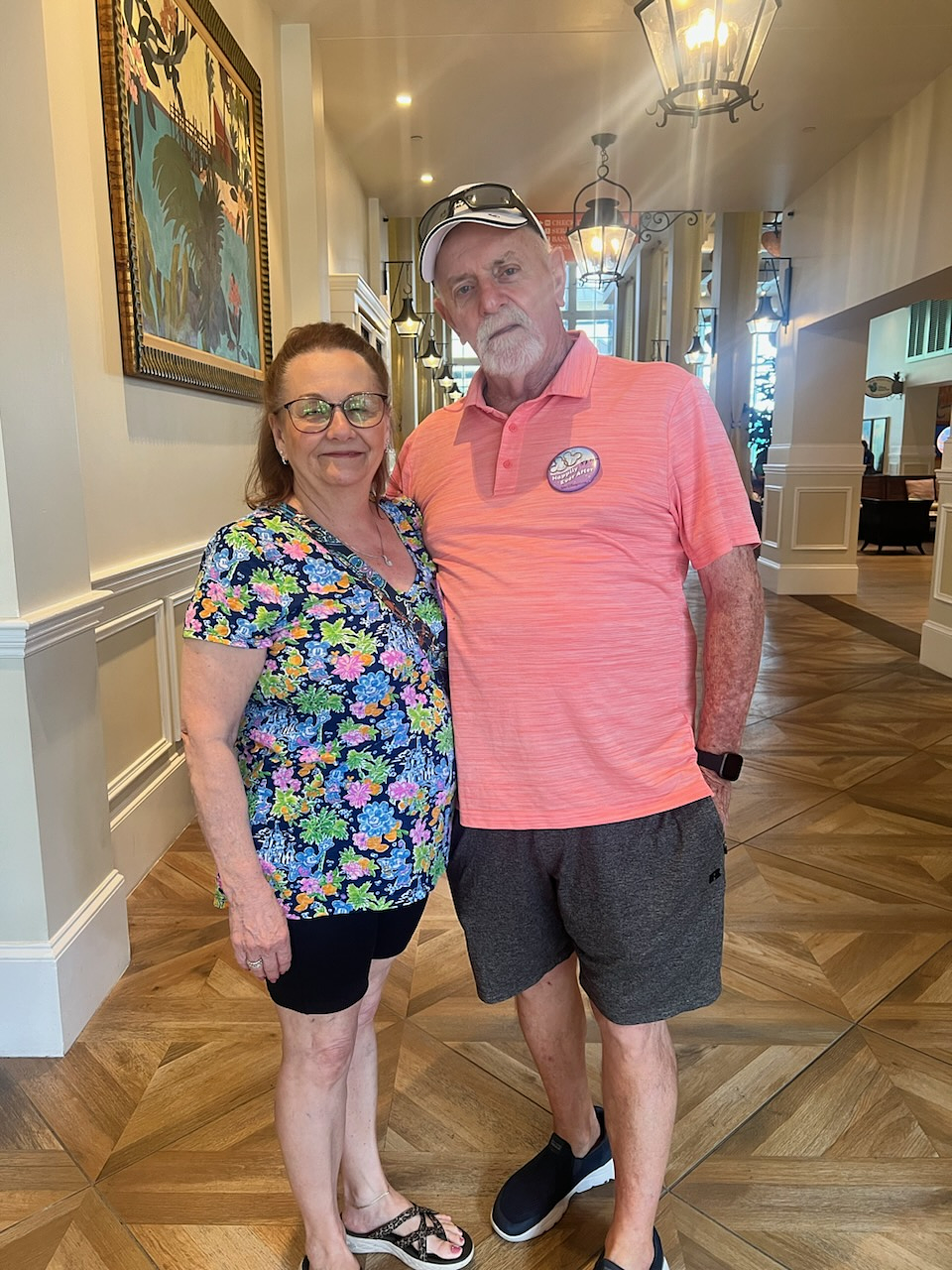 A smiling older couple stands arm-in-arm in a beautifully decorated hallway at Disney World, with warm lighting, wooden floors, and hanging lanterns. The woman wears a colorful floral top and black shorts, and the man wears a coral polo shirt, dark shorts, a white cap, and a celebratory button. The photo was taken during their anniversary celebration trip