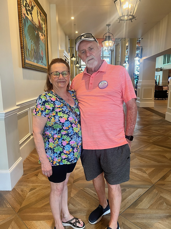 A smiling older couple stands arm-in-arm in a beautifully decorated hallway at Disney World, with warm lighting, wooden floors, and hanging lanterns. The woman wears a colorful floral top and black shorts, and the man wears a coral polo shirt, dark shorts, a white cap, and a celebratory button. The photo was taken during their anniversary celebration trip
