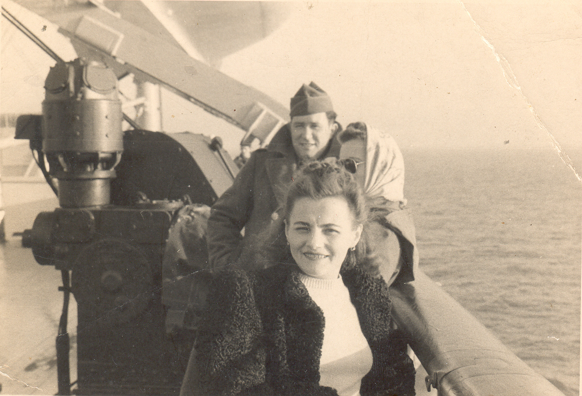 A young woman smiles on the deck of a ship, wearing a dark coat over a light sweater. A man in military uniform and another woman stand behind her, with the ocean stretching out in the background.