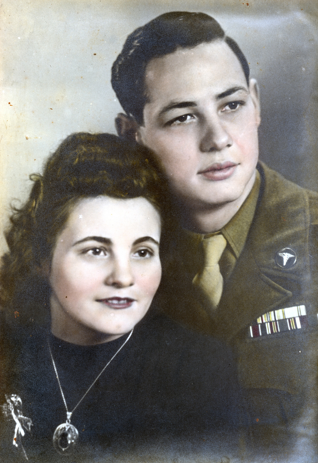 A colorized portrait of a young couple from the 1940s. The woman has dark curled hair and wears a black dress with a necklace and brooch. The man, in a military uniform with medals and a medical insignia, stands slightly behind her, looking off into the distance.