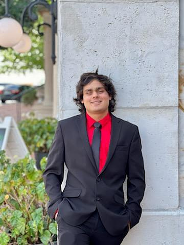 A young man celebrating his 21st birthday stands confidently in a black suit, red shirt, and black tie, leaning against a light gray stone wall. He smiles softly with his hands in his pockets, and decorative lights and greenery appear in the background.