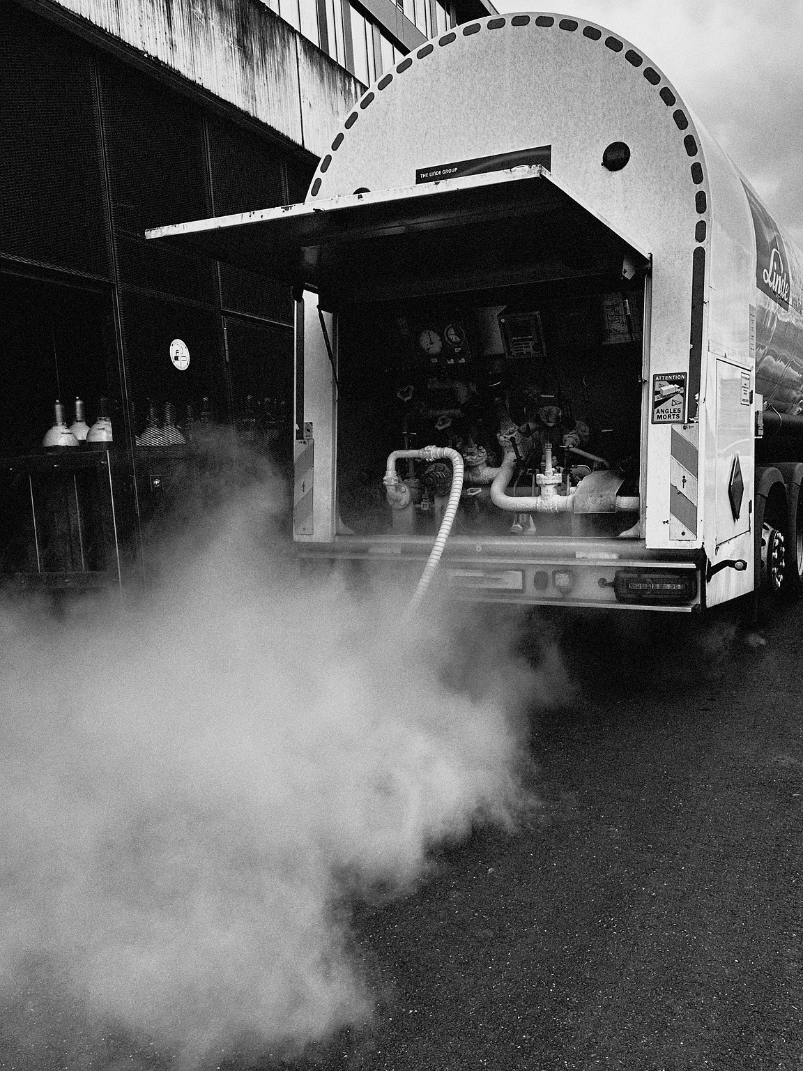 A black and white photograph of the rear of a tanker truck with its compartment open, exposing various gauges and pipes. There is a large plume of nitrogen billowing from the equipment, spreading across the ground. The truck is parked near a building with visible pipes and tanks alongside it. The atmosphere appears industrial.