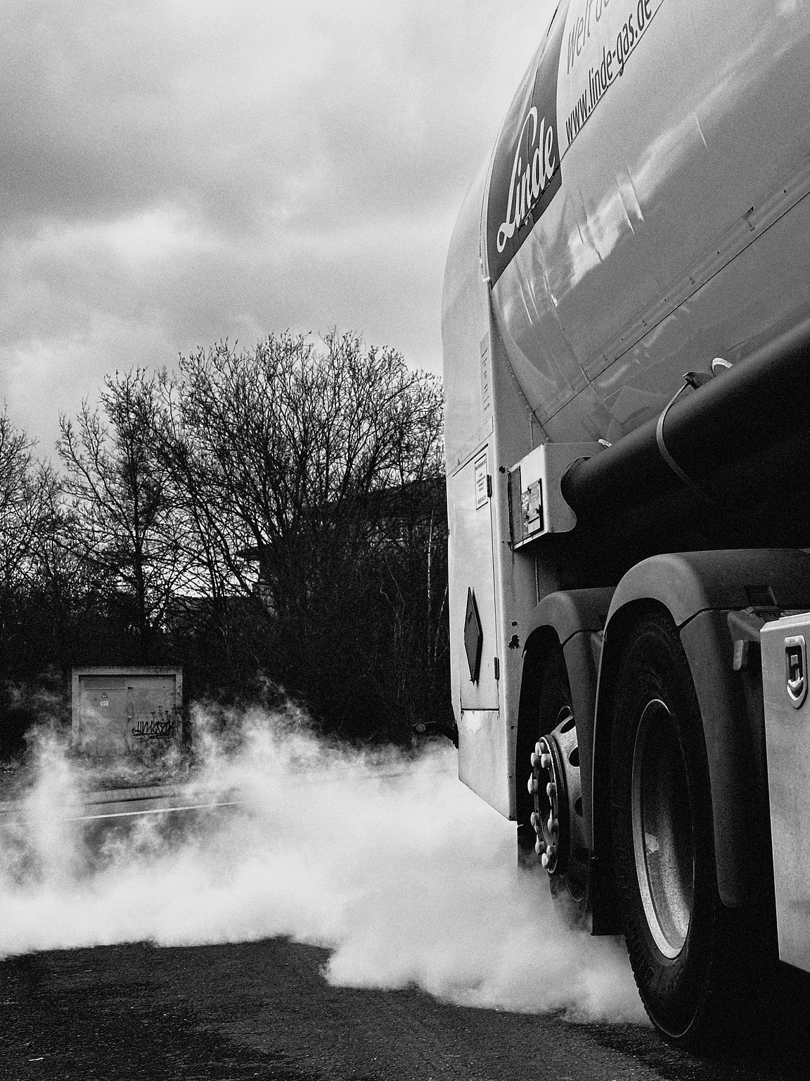 A black and white image of a tanker truck with the logo "Linde" on its side, parked on a road. The truck is releasing a cloud of nitrogen near its rear wheels. In the background, there are leafless trees and a small structure with graffiti. The sky is overcast.