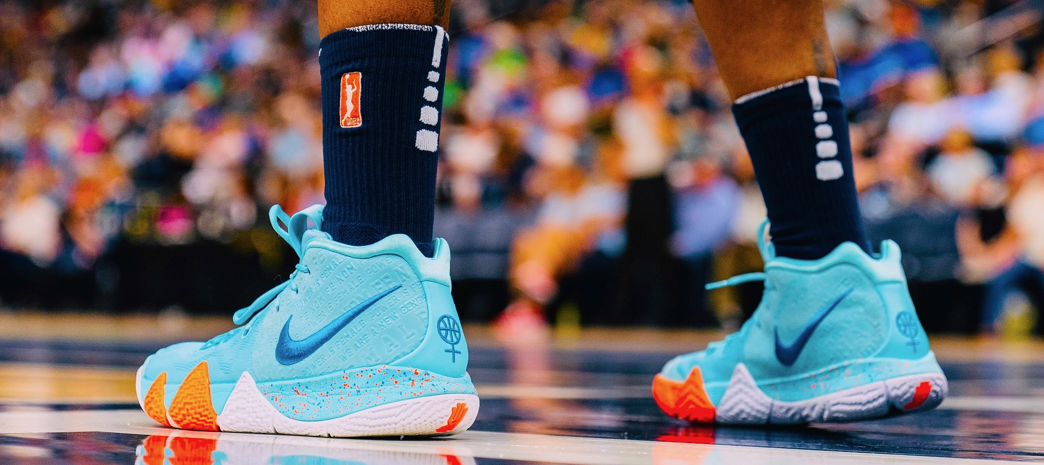 A player stands on a basketball court. She's shown only from the knees down: her feet in two light blue and orange basketball shoes with the nike swoosh and tall navy blue socks with the orange WNBA logo on one side. In the background are the pixelated faces of fans. 